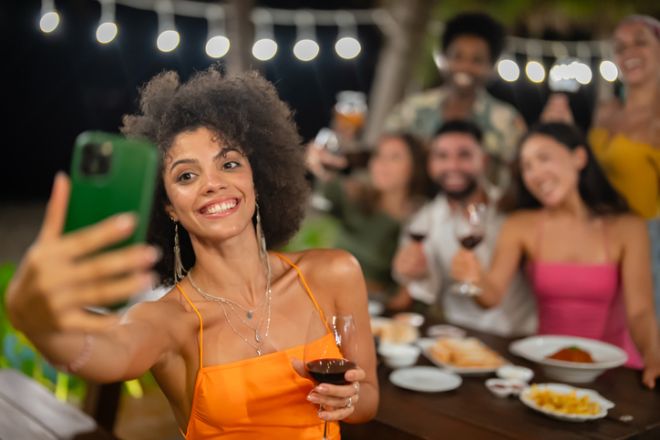 Young woman taking a cheerful selfie with friends during an outdoor dinner party at night, all raising wine glasses and enjoying the festive atmosphere