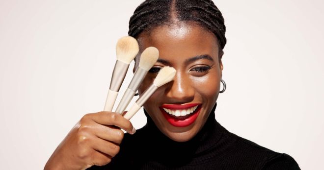 Studio, makeover and woman with makeup brush, portrait and equipment for beauty, happy and selfcare. White background, black person and cosmetic for self expression, creative and tools for foundation