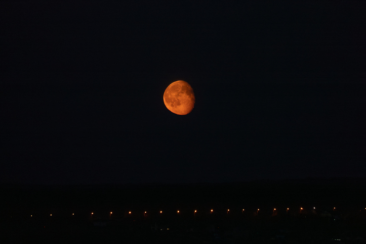 Full orange moon illuminating the dark night sky, glowing above a distant line of warm city streetlights creating a striking nocturnal astronomical view