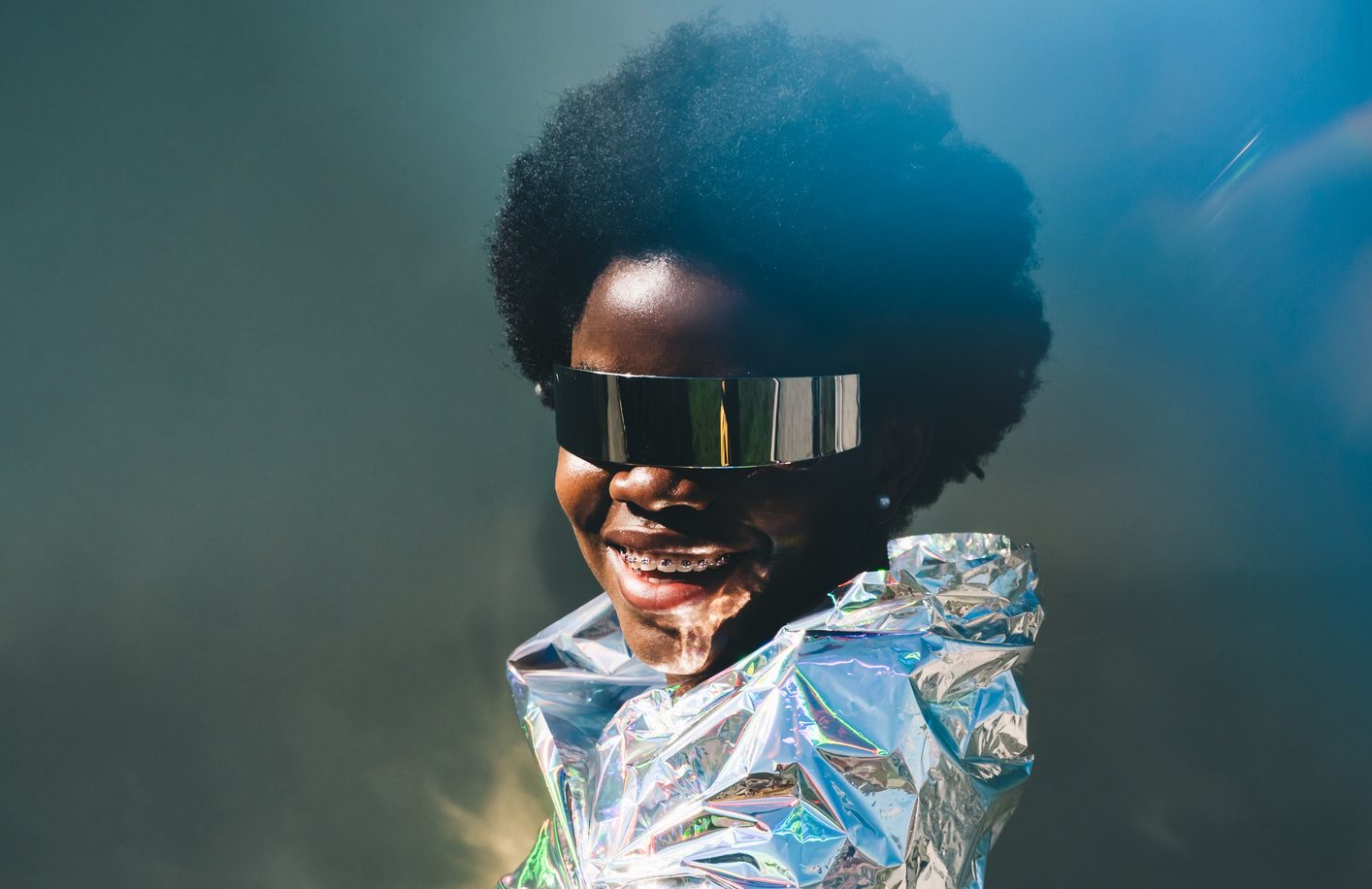 A futuristic portrait of a smiling African American woman wearing reflective visor sunglasses and wrapped in a metallic cloak.