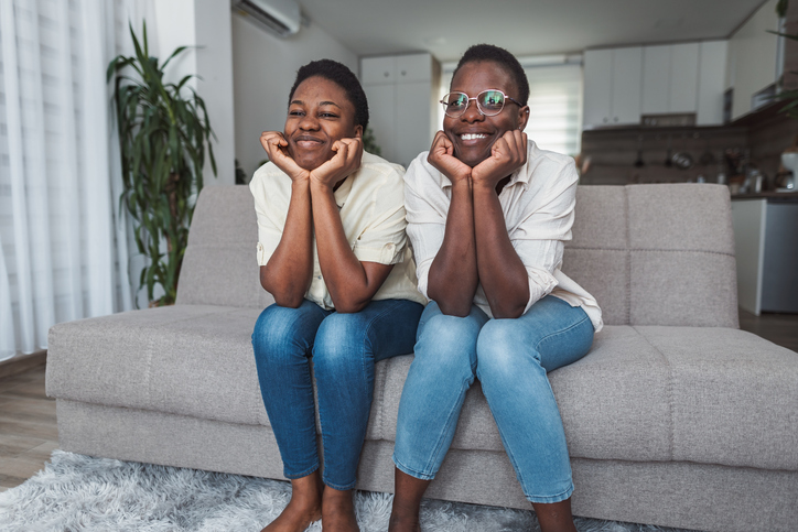 Two cheerful black twin sisters watching television sitting on sofa at home