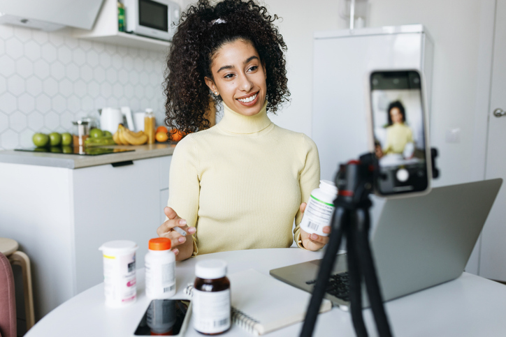 Selective focus on black female blogger nutritionist holding bottle of food supplement sitting at kitchen table in front of smartphone camera mounted on tripod, giving advice or tips to stay healthy