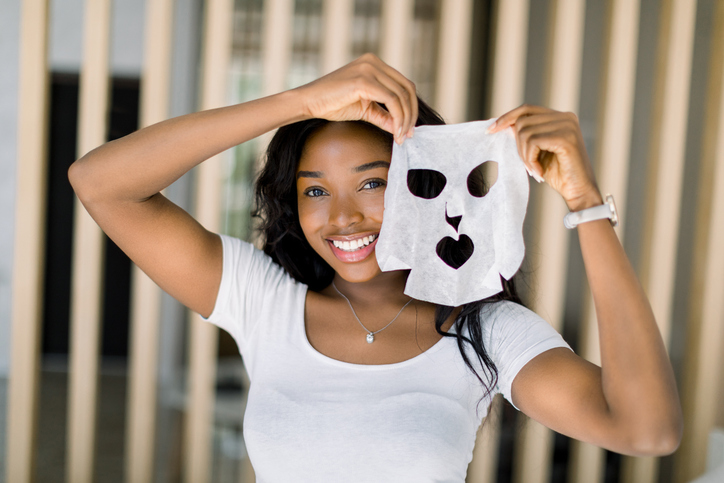 Beautiful young African American lady in white t-shirt, enjoying her beauty procedures at home, going to apply a white face sheet mask on face. Spa procedures at home